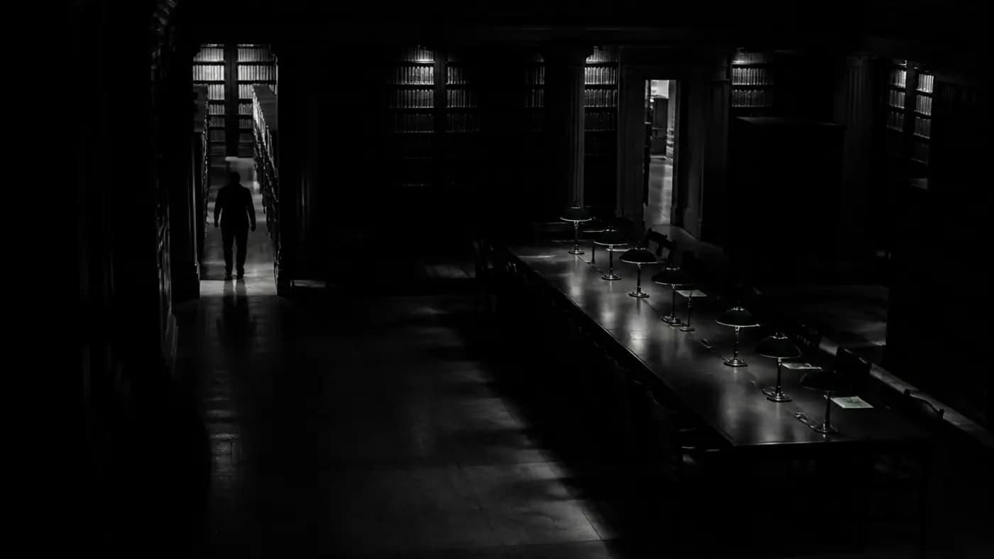 A law library reading room after hours, lamps lit along an empty table, one figure between the stacks.